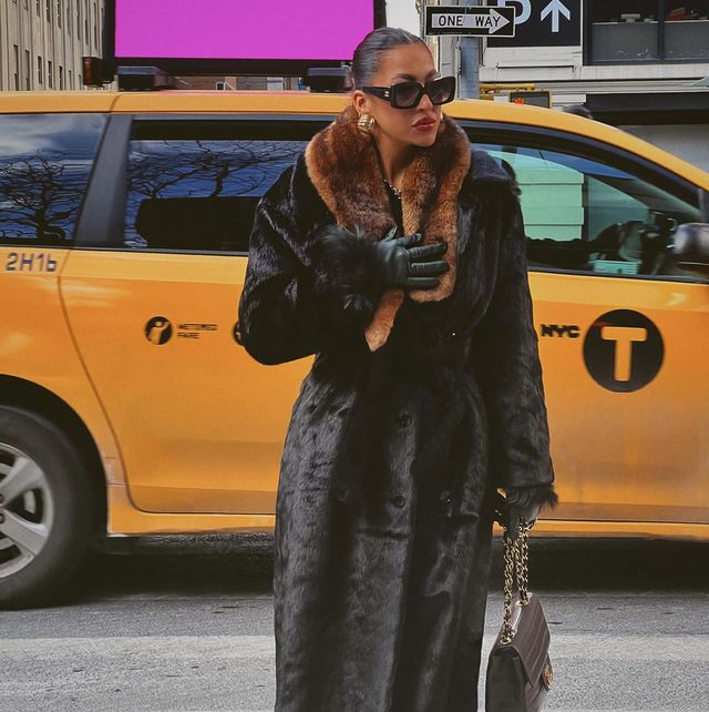 a woman wearing a fur oat crossing the street in front of a yellow taxi in New York City